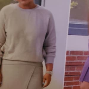 May include: A woman wearing a light gray sweatshirt and a matching wrap skirt. She is wearing a watch and hoop earrings. The background includes a brick wall and a poster with the text "Simplicity".