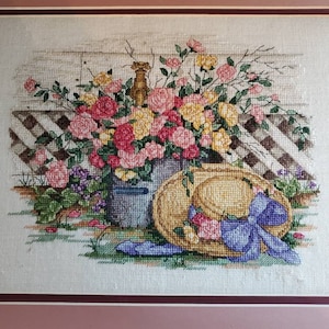 May include: A cross-stitch embroidery featuring a bouquet of pink and yellow roses in a grey container, placed next to a straw hat with a blue ribbon. The background shows a white lattice fence, all framed within a wooden border with matting.