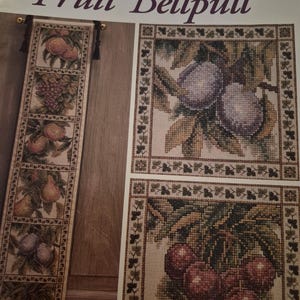 May include: A cross-stitch pattern titled "Fruit Bellpull" showing detailed images of various fruits. The design includes plums, pears, grapes, and cherries, with a decorative border. The colour palette includes shades of brown, green, purple, and red.