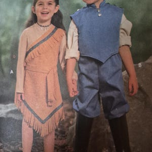May include: A tan sleeveless dress with fringe trim and a blue patterned border, worn over a long-sleeved beige shirt. Beside it, a blue vest with silver buttons, a beige long-sleeved shirt, blue trousers, and black knee-high boots.