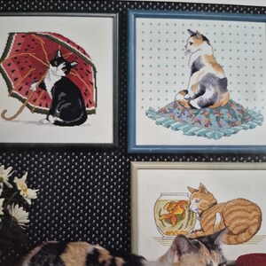 May include: Three framed cross-stitch pictures featuring cats. One shows a black and white cat with a watermelon umbrella, another a calico cat on a floral cushion, and the third an orange tabby cat beside a goldfish bowl. A calico cat sits below.