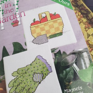 May include: Cross-stitch magnets featuring garden-themed designs. One magnet shows a basket of red tomatoes with a trowel, and the other depicts a green and purple gardening glove. The packaging reads "In the Garden" and "Exclusively Ours."
