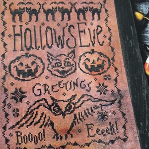 May include: Cross-stitch artwork in a black frame, celebrating Halloween. The design includes black cats, pumpkins, and a bat, with the text "Hallow's Eve Greetings" and spooky phrases. The background is a mottled orange and brown.