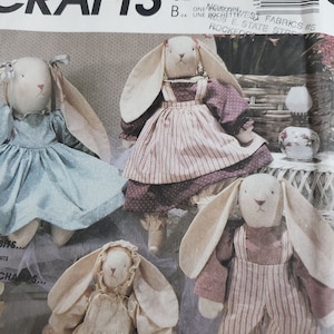 Op de afbeelding: Een McCalls Crafts naaipatroon-envelop met vier stoffen konijnenpoppen. De konijnen dragen verschillende outfits, waaronder een lichtblauwe jurk, een paarse jurk met een gestreept schort, een crèmekleurige jurk met een mutsje en een gestreepte tuinbroek.