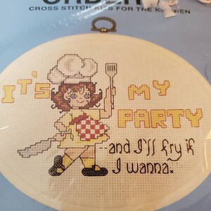 May include: A cross-stitch kit featuring a cartoon chef holding a spatula and a checkered heart. The text reads "It's My Party and I'll fry if I wanna." The kit is in a round frame with a metal loop for hanging.