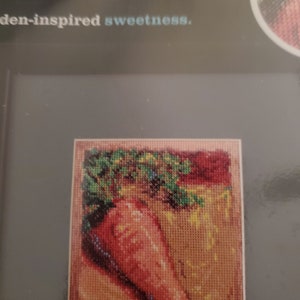 May include: Cross-stitch pattern of a carrot with green leaves on a beige background. The text "Carrot" is above the image and the text "Garden-inspired sweetness." is below the image.