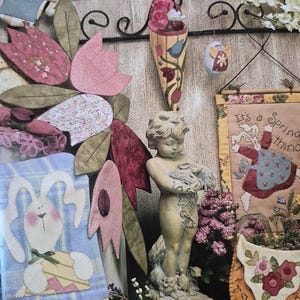 Op de afbeelding: Een lentethema-display met een stenen cherubijnbeeld dat vogels vasthoudt, omringd door textielkunst. Bevat een konijn, bloemaccenten en een banner met de tekst "It's a Spring thing". Het kleurenpalet omvat roze, groen en blauw.