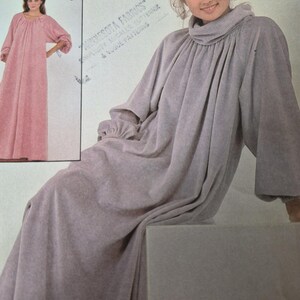 Può includere: Una busta per cartamodello che presenta un abito a maniche lunghe fino ai piedi con scollo ad anello arricciato e polsini elasticizzati. L'immagine principale mostra l'abito in color lavanda chiaro, mentre un riquadro mostra una versione rosa etichettata B.
