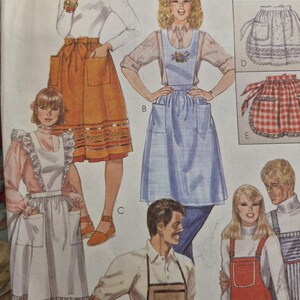May include: A McCall's sewing pattern envelope featuring illustrations of various apron styles. Designs include a mustard yellow apron with pockets, a blue pinafore with floral embroidery, a white apron with lace trim, and red checkered aprons.