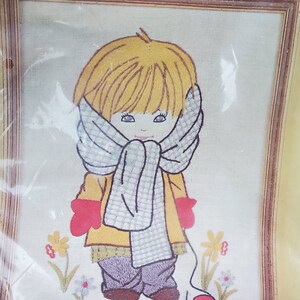 May include: Embroidered artwork of a child wearing a scarf, coat, and mittens, with the text "READY FOR ANYTHING!". The child has blonde hair and is surrounded by embroidered flowers. The artwork is framed in a wooden frame.