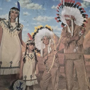 May include: A Simplicity sewing pattern envelope featuring four individuals wearing tan fringe costumes. The group includes two people in dresses with silver accents and two in pants and shirts with feathered headdresses, one holding a bow.