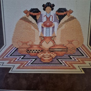 May include: A cross-stitch embroidery depicting three figures with dark hair and tan garments surrounding a collection of terracotta-colored pottery. The figures are positioned on a geometric, multi-colored patterned base with a dark brown border.