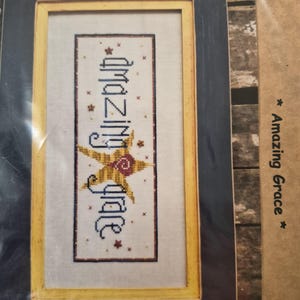 Op de afbeelding: Ingeklemd kruissteekkunstwerk met de woorden "amazing grace" in blauwe draad. Het ontwerp bevat een gele bloem en rode accenten tegen een crèmekleurige achtergrond. De lijst is een combinatie van geel en donkerblauw.