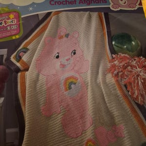 Puede incluir: Una manta afgana de Care Bears tejida a crochet en crema, rosa y colores arcoíris. La manta presenta un gran personaje de Care Bear rosa con un arcoíris en su vientre y las palabras "Care Bear" en rosa. El borde tiene rayas naranjas y moradas.