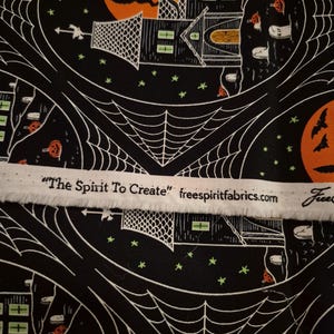 Op de afbeelding: Zwarte stof met een herhalend Halloween-patroon van spookhuizen, oranje manen, vleermuizen en spinnenwebben. Een witte zelfkant toont de tekst "The Spirit To Create" en freespiritfabrics.com in zwarte letters.