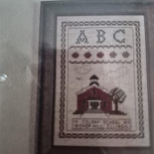 May include: A framed cross-stitch sampler featuring the letters A, B, and C at the top, a row of five red shapes, and a depiction of a red schoolhouse with a tree. The text at the bottom reads A COLONY SCHOOL IN BISHOP HILL ILLINOIS.
