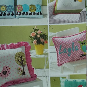 May include: Simplicity pattern showing several cushion designs. The cushions feature appliqued motifs such as a heart and bee, an owl, a dragon, and abstract shapes. The cushions are in various colours, including pink, blue, green, and white.