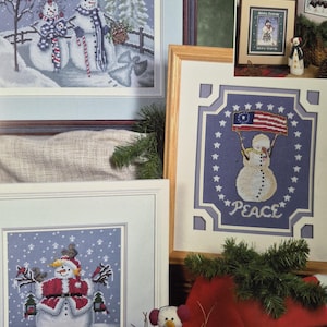 Könnte beinhalten: Gerahmte Kreuzstichkunst mit Schneemännern in Winterszenen. Ein Stück zeigt Schneemänner mit einem Zaun und Bäumen, ein anderes hat einen Schneemann, der eine Flagge mit dem Wort "PEACE" hält, und ein drittes hat einen Schneemann mit dem Text "Joy to the World".