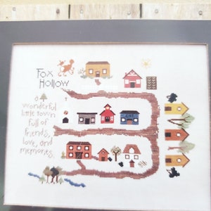 May include: Cross-stitch sampler titled "Fox Hollow" featuring a charming village scene. The design includes various colorful houses, trees, and a winding path, with text that reads "a wonderful little town full of friends, love, and memories."