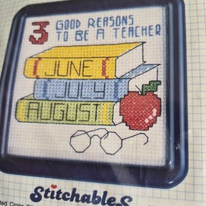 May include: A WiseGuys Stitchables cross stitch kit featuring a design with three stacked books labeled June, July, and August, a red apple, and eyeglasses. The text reads 3 Good Reasons To Be A Teacher. The kit includes a blue frame and materials.