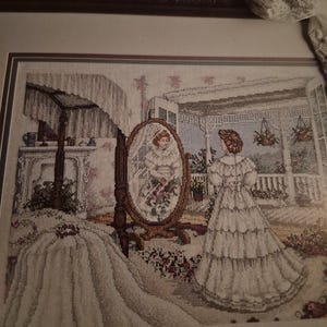 May include: A framed cross-stitch embroidery depicting a person in a long white dress standing before an oval floor mirror. The scene includes a canopy bed, a porch with hanging plants, and the word Forward printed at the top of the frame.