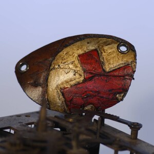 May include: Close-up of a distressed, abstract object in brown, red, and white. The object, possibly leather, has two holes and sits on a metal structure. The colour palette is muted, with signs of wear and tear.