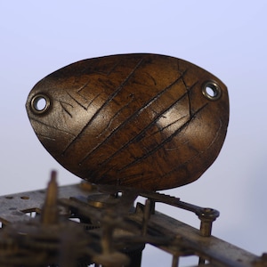 Pode incluir: Um close-up de um mostrador de relógio de madeira marrom com dois anéis de metal. O mostrador é esculpido com linhas e tem uma aparência desgastada e envelhecida.