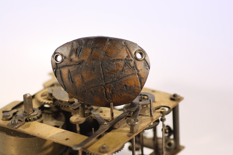 May include: A brown, textured leather piece with two metal eyelets, possibly a watch component, sits atop a complex arrangement of clock gears and mechanisms. The leather has a worn appearance, suggesting age or use.