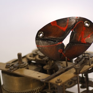 Pode incluir: Um close-up de um mecanismo de relógio vintage com uma peça de couro em forma de peixe vermelho e preto presa ao topo. O peixe tem uma rachadura no meio e dois pequenos furos no lado.