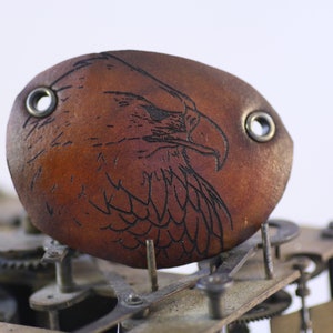 Può includere: Una benda sull'occhio in pelle marrone con un'incisione nera dettagliata di un'aquila. La benda ha due occhielli metallici ed è posta su uno sfondo di ingranaggi di orologio, suggerendo un'estetica steampunk.
