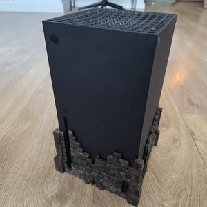 Puede incluir: Una consola de juegos negra colocada verticalmente sobre una base decorativa de ladrillos gris oscuro. La consola tiene una parte superior perforada y un pequeño botón circular. La base añade una estética única.