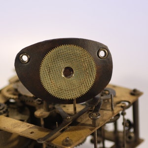 May include: Close-up of a vintage clock mechanism. A dark brown, leather-like piece with a circular, textured, brass-colored component is centered. The mechanism features various gears and metal components, suggesting a repair or restoration project.