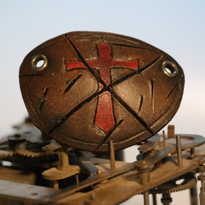 Puede incluir: Un parche para el ojo de cuero marrón con un diseño de cruz roja. El parche para el ojo está hecho de varias piezas de cuero que están cosidas juntas. El parche para el ojo está sentado en un mecanismo de engranaje de metal.