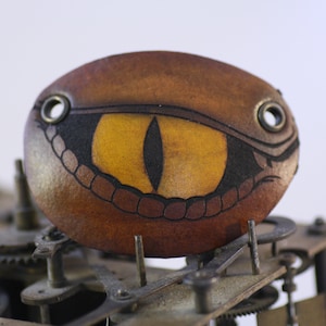 May include: A brown leather eye-shaped embellishment with a yellow iris and black detailing. The eye has a stylized design with a snake-like appearance. Two metal grommets are on either side of the eye. The object is set against a background of clock gears.