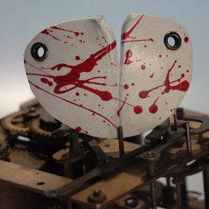 May include: A white, abstract mask with red paint splatters, featuring two eye holes. The mask is positioned above the intricate gears and mechanisms of a clock.