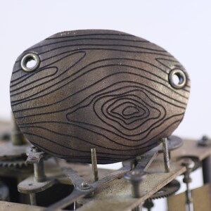 May include: A close-up of a brown, oval clock component with a wood grain design. The component has two metal eyelets and is part of a clock mechanism with gears and metal parts. The background is white.