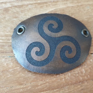 Pode incluir: Remendo oval de couro castanho com um símbolo triskel preto. O remendo tem dois ilhós de metal e é provavelmente para roupas ou acessórios. O couro tem um aspeto ligeiramente envelhecido. Mede cerca de 8 cm de comprimento.