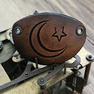 May include: A brown leather cuff with a crescent moon and star design. The cuff has two holes for attaching to a belt or other item.