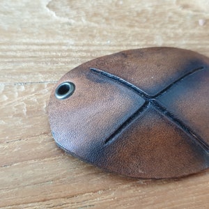 May include: Oval-shaped brown leather eye patch with an X-shaped design and two metal eyelets. The leather has a distressed look with darker edges, suggesting a vintage or rustic aesthetic. The eye patch is designed to be worn over one eye.