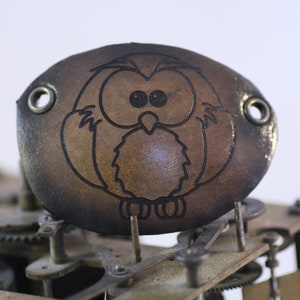 May include: A brown leather patch featuring a black owl design. The oval-shaped patch has two metal eyelets. The owl is centrally placed, with large eyes and a simple outline. The patch is set against a background of clockwork mechanisms.