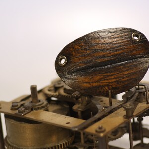 May include: Close-up of a vintage clock mechanism with a wooden oval-shaped piece on top. The clockwork is made of brass and has intricate gears and levers.