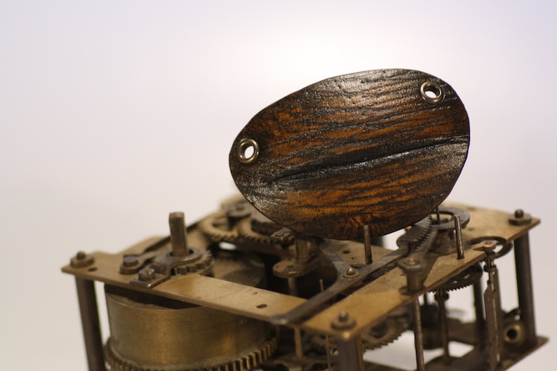 May include: Close-up of a vintage clock mechanism with a wooden oval-shaped piece on top. The clockwork is made of brass and has intricate gears and levers.