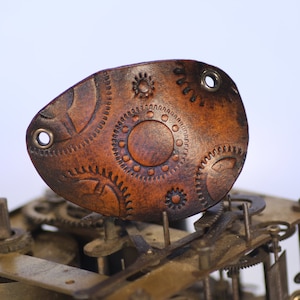May include: A brown leather patch with a steampunk design featuring gears and circles. The patch has two holes punched in it.