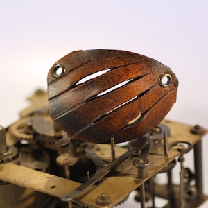 May include: A brown leather clock face with four cutouts and brass rivets. The face is mounted on a brass clock mechanism with gears.