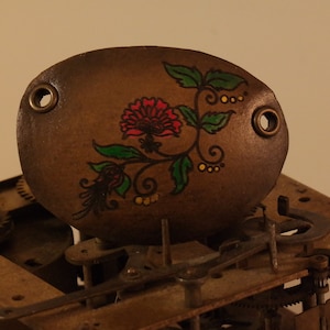 May include: A close-up of a brown, oval-shaped metal plate with a painted floral design. The plate has two holes on either side. The design features a red flower with green leaves and yellow accents.