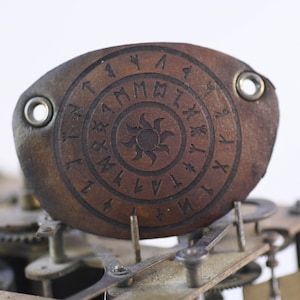 Puede incluir: Un parche de ojo de cuero marrón con runas grabadas y un símbolo de sol. El parche tiene dos ojales metálicos y está colocado sobre un fondo de engranajes y mecanismos de relojería. El diseño es circular y detallado.