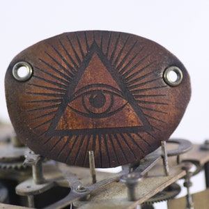 Può includere: Toppa ovale in pelle marrone con un disegno inciso al laser di un occhio all'interno di un triangolo, linee radianti e occhielli metallici. La toppa è posta su uno sfondo di ingranaggi di orologeria.