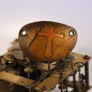 Könnte beinhalten: Ein braunes Lederarmband mit einem Kupferkreuz-Design. Das Armband hat zwei Metallösen an den Seiten.