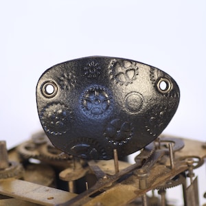 May include: A black metal clock face with a decorative design of gears and circles. The face is attached to a clock mechanism with visible gears.