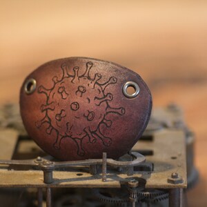 Op de afbeelding: Een bruin leren ooglapje met een zwart virusontwerp en metalen oogjes. Het ovale ooglapje ligt op een vintage uurwerk, met een houten achtergrond.